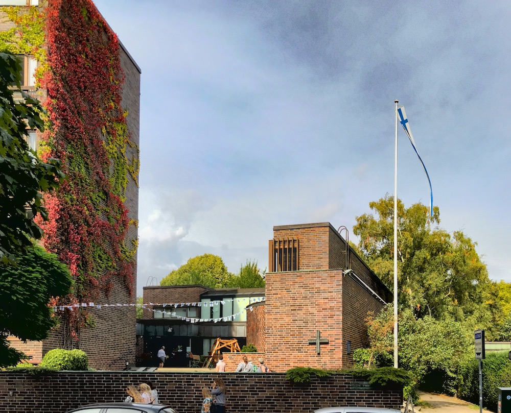 Die Fassade der Finnischen Seemannskirche: ein eher erckiges Gebäude aus rotem Backstein, davor ein Platz und links die hohe Backteinfassade mit herbstlich gefärbten Blättern
